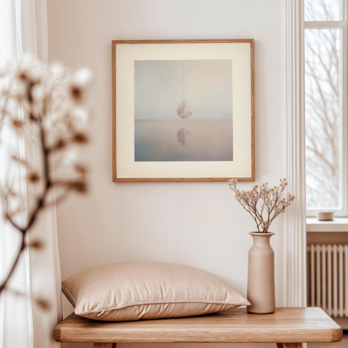 Framed print of white feather with soft colours hanging above a beige cushion on a wooden side table