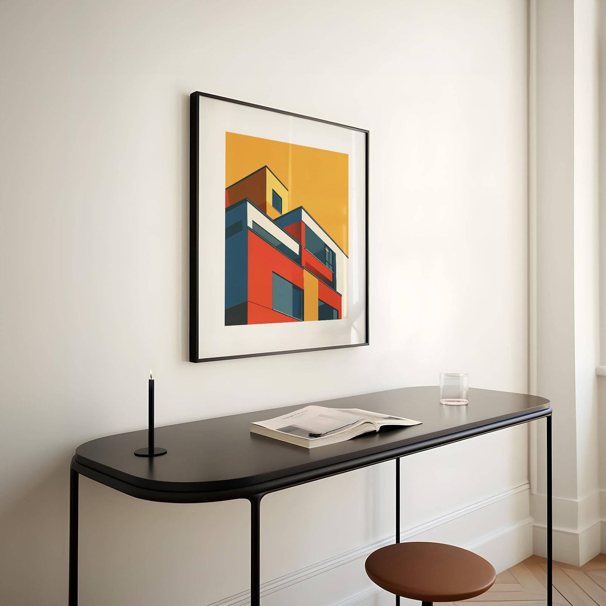 Framed print of modernist building illustration in bright colours above a black desk with a candle and book