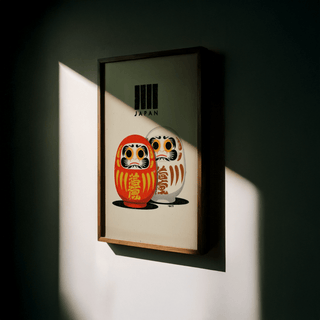 Framed Daruma print showing red and white dolls with Japan text in a shadowed room