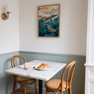 Whale Art Print on a wall above a small dining table with two wooden chairs and croissant