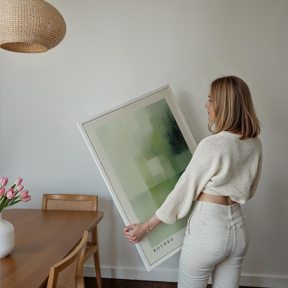 Framed print with green abstract shapes held by a woman with a white jumper in a dining area