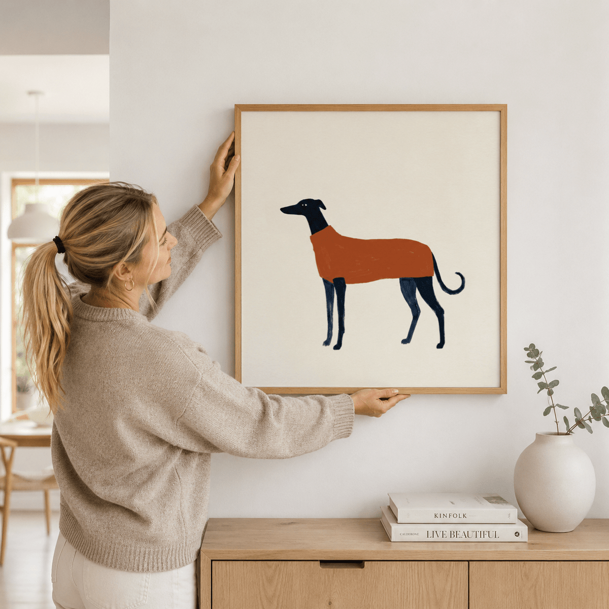 Greyhound art print held by woman above sideboard with plant and books in natural tones