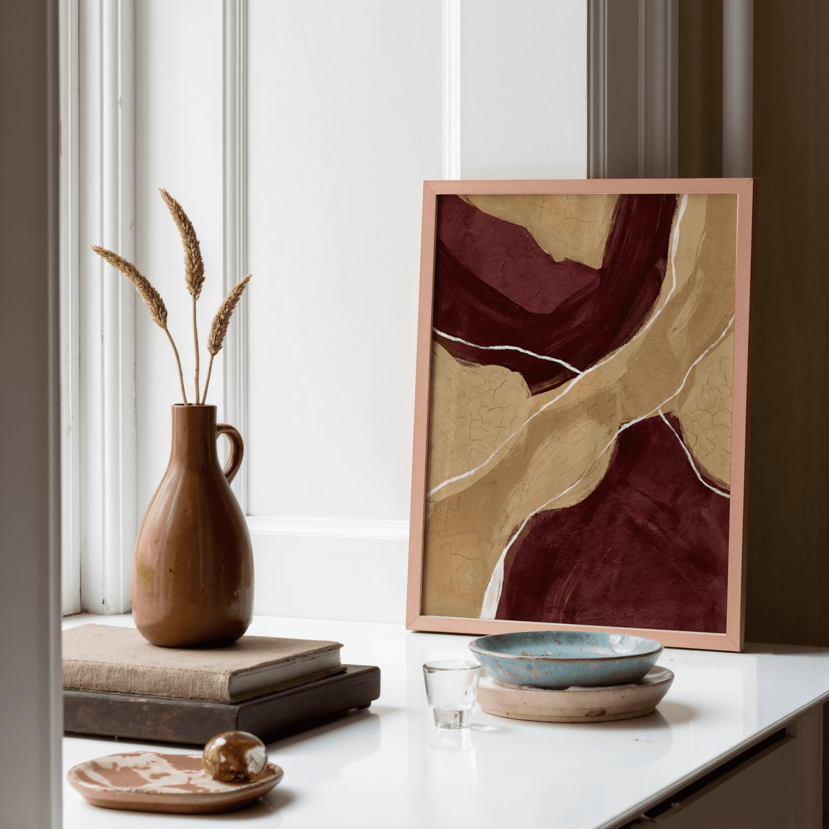 Framed print of neutral organic abstract art with beige and burgundy shapes next to a brown vase with dried grasses