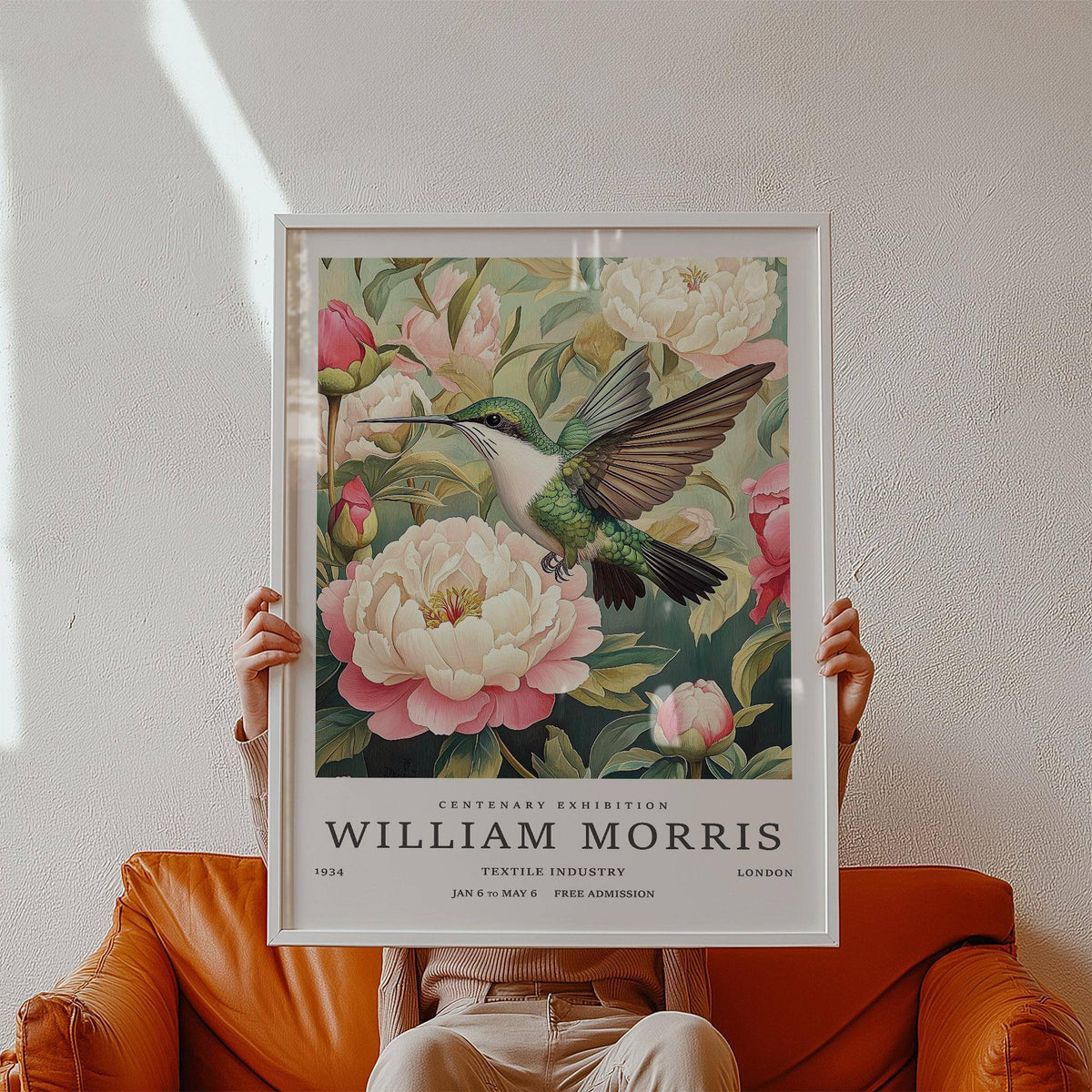 Framed print of hummingbird and peonies botanical illustration held sitting on a brown leather sofa