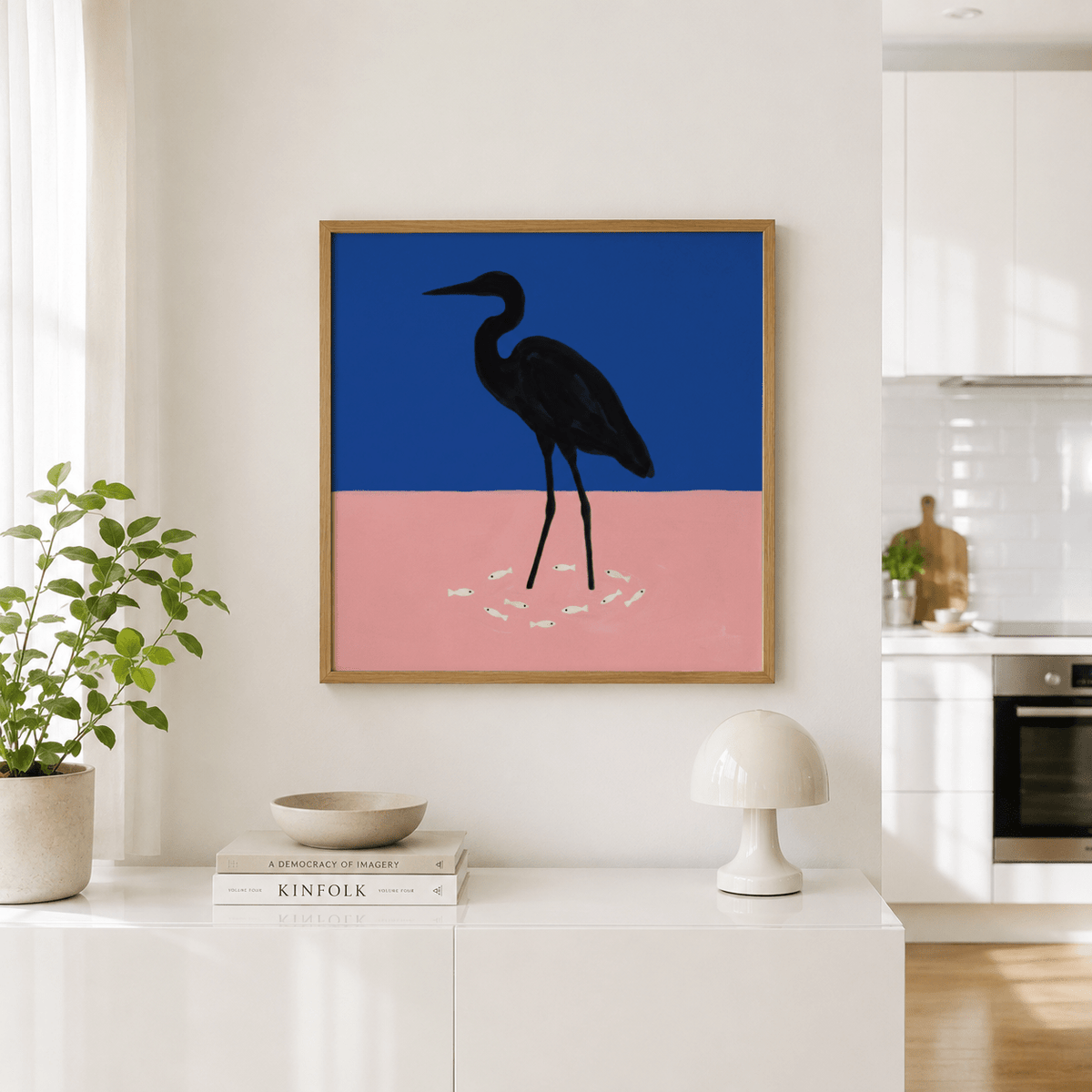 Heron art print with a black heron silhouette on blue and pink background above a white sideboard with books and a lamp