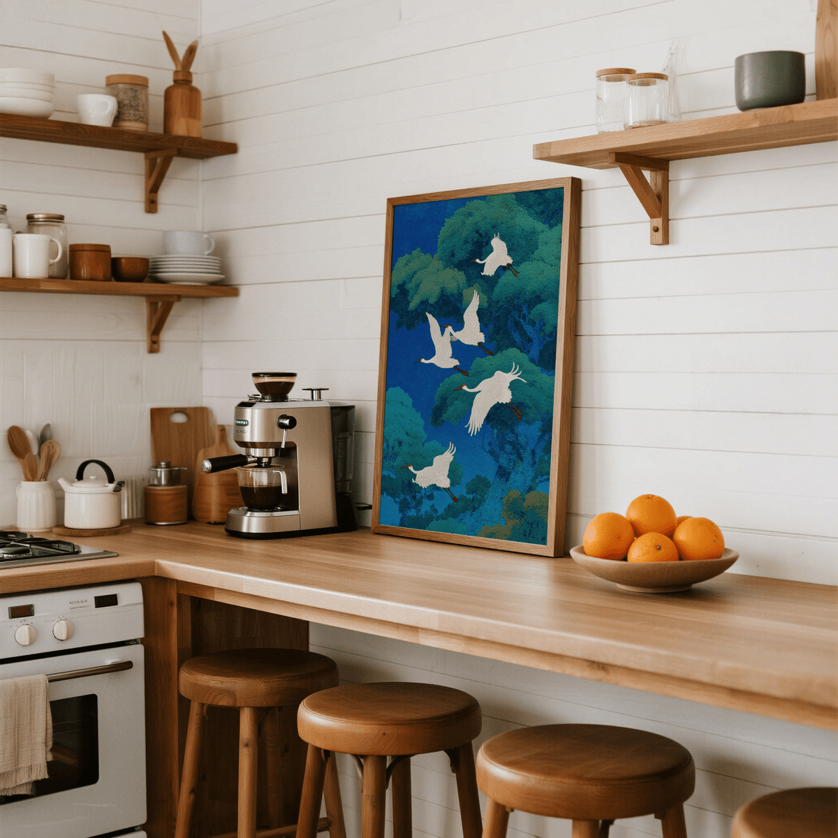 Print leaning on a kitchen counter with white cranes flying among green trees in a blue background and a bowl of oranges