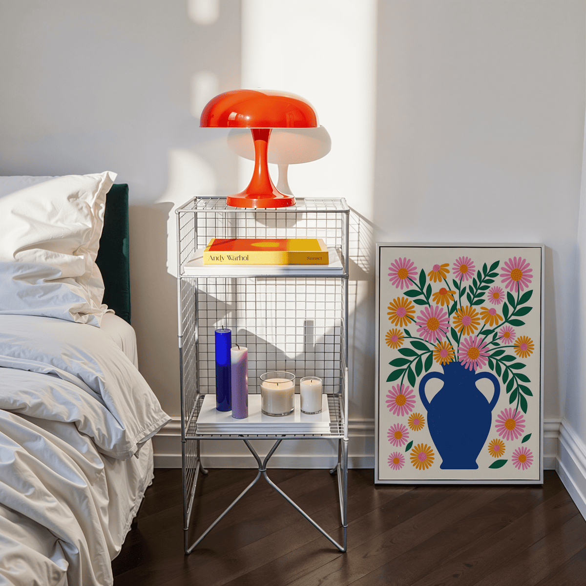 Print leaning on the floor of floral vase with orange and pink flowers beside a bed and orange lamp