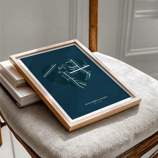Framed Love Map Poster with a heart-shaped map in blue tones leaning on a beige fabric chair with books