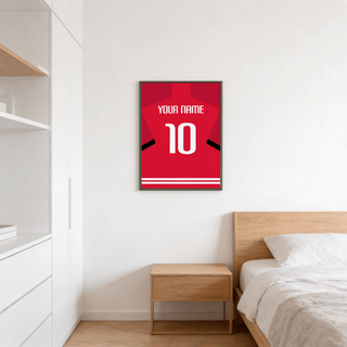 Print on a wall in a bedroom of a red football shirt poster with black sleeve stripes and white text above a bedside table
