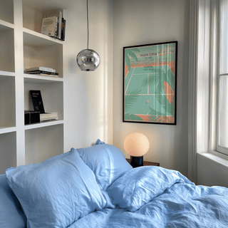 Print on a wall in a bedroom with blue bedding and a round lamp featuring a tennis court with palm trees in pastel colours