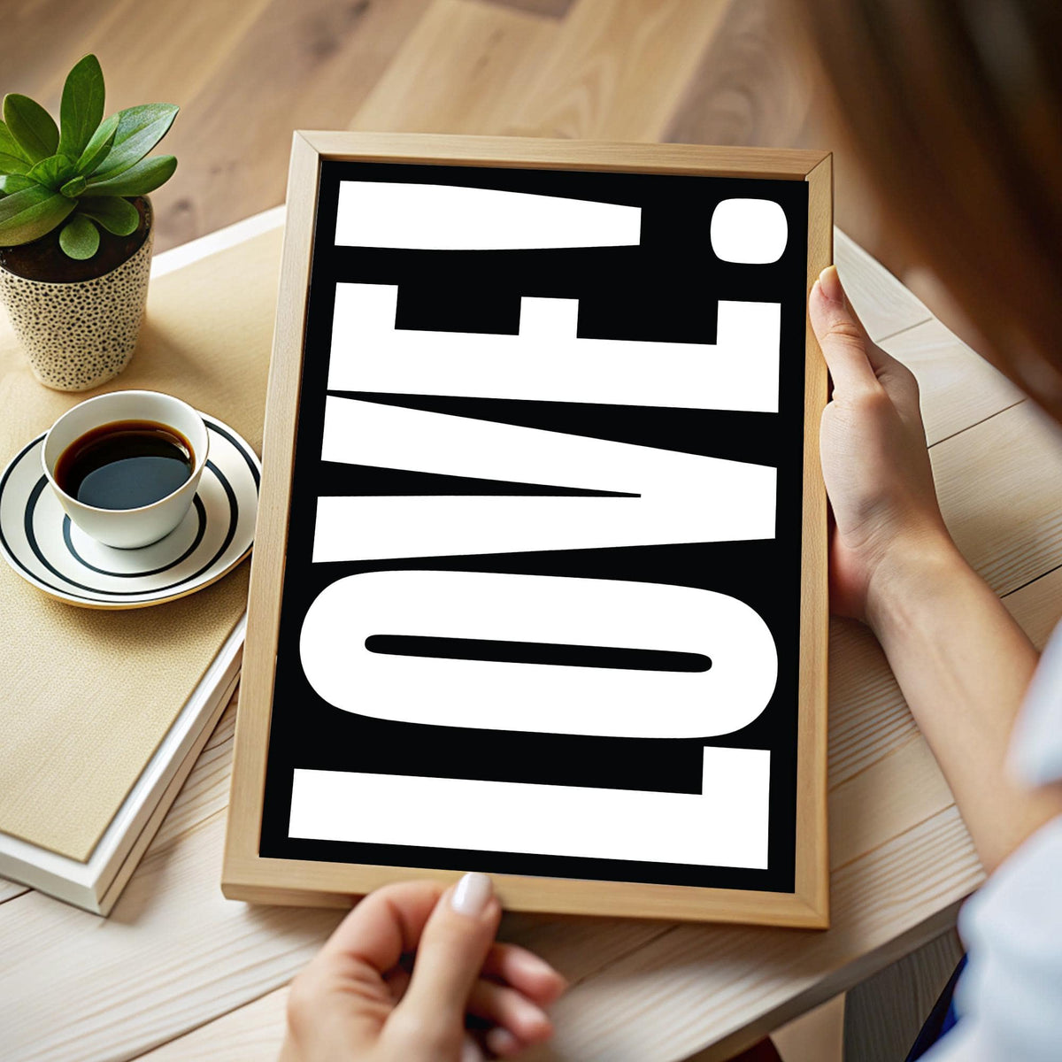 Framed print close-up of LOVE typography in white on black held above a wooden table with a plant and coffee