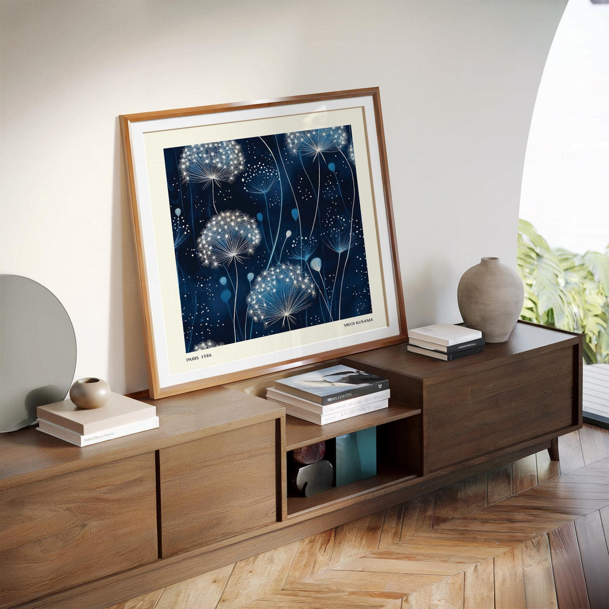 Blue art work of minimalist dandelion seed heads leaning on a wooden sideboard next to a vase