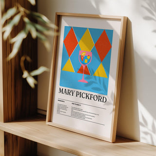 Framed print of Mary Pickford cocktail illustration in red yellow and blue leaning on a surface with a plant