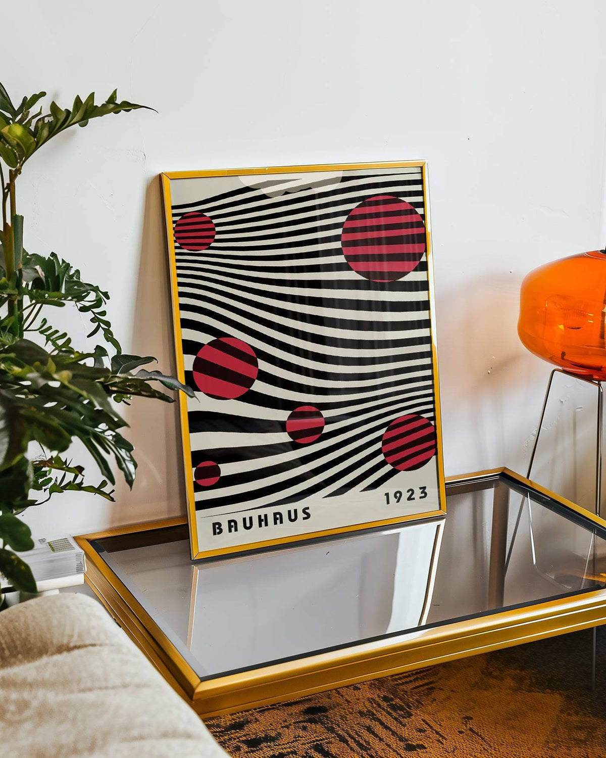 Large Bauhaus wall art with red striped circles on wavy black lines in a gold frame leaning on a glass table with an orange lamp