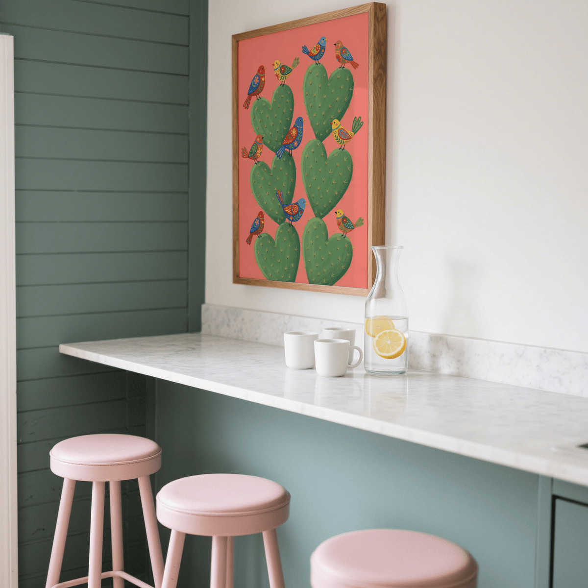 Print on a wall in a kitchen showing Cactus Heart Poster with green and orange folk-art style and birds above pink stools