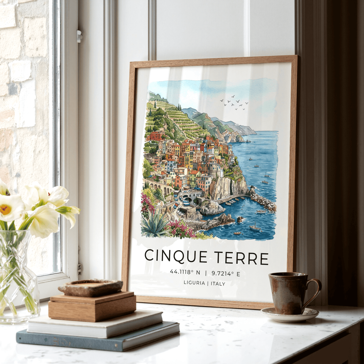 Print leaning on a windowsill featuring a colourful coastal village with blue sea and green hills alongside flower vase and cup