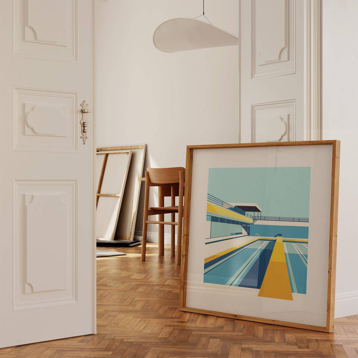 Print leaning on floor of a minimalist room showing an urban swimming pool in blue and yellow tones with a chair nearby