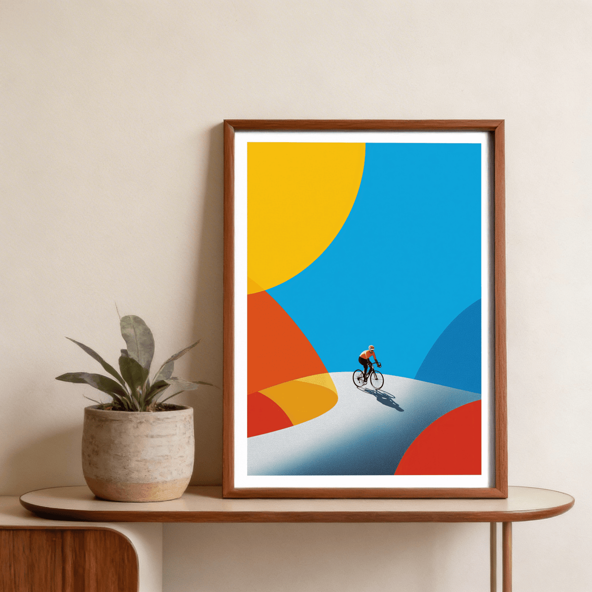 Framed Cycling Art Print of a cyclist on a winding road with bright colour shapes on a shelf with a plant