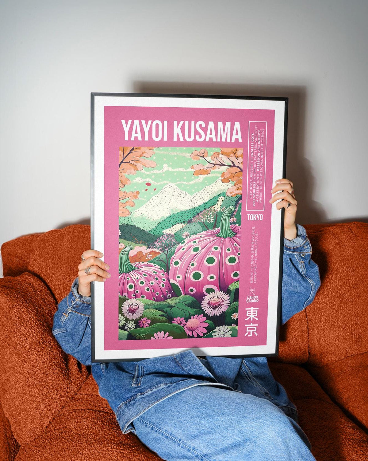 Wall Art with Pink polka dot pumpkins and green hills held by person on rust sofa