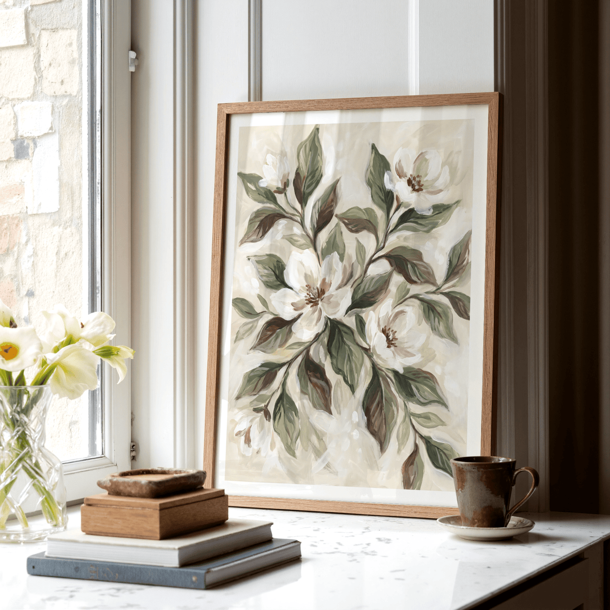 Magnolia wall decor print leaning on a windowsill with brown leaves and cream flowers near a brown cup