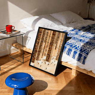 Print leaning on floor of library art print with beige and brown book stacks in a bedroom with blue patterned blanket