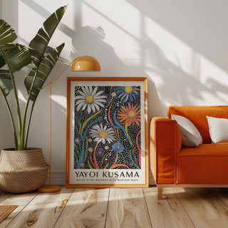 Print leaning on a floor beside an orange sofa and a tall plant with yellow lamp above