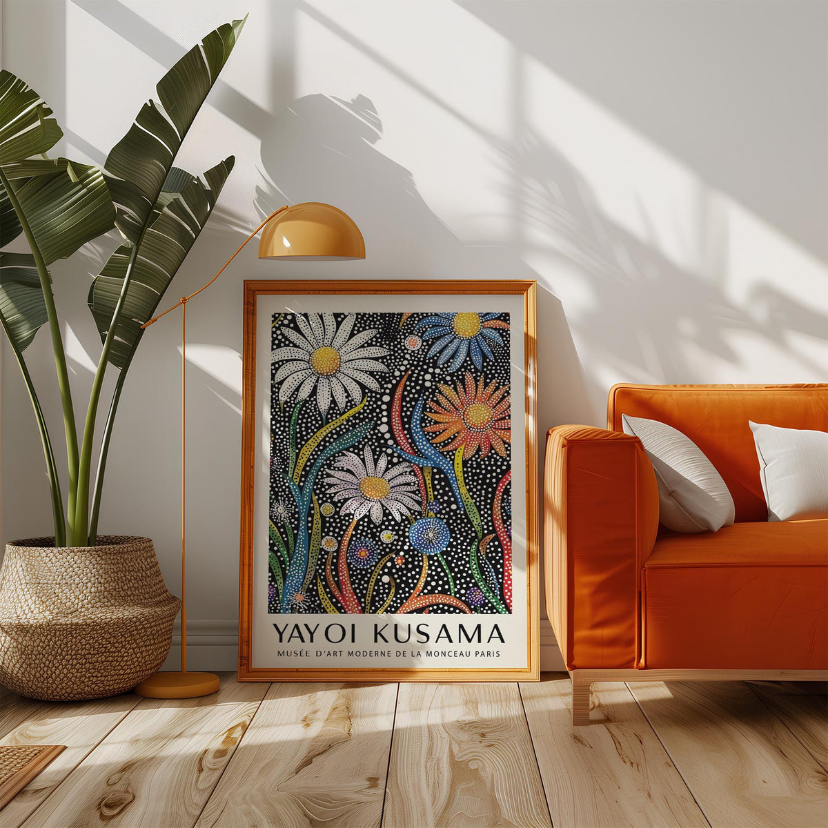 Print leaning on a floor beside an orange sofa and a tall plant with yellow lamp above