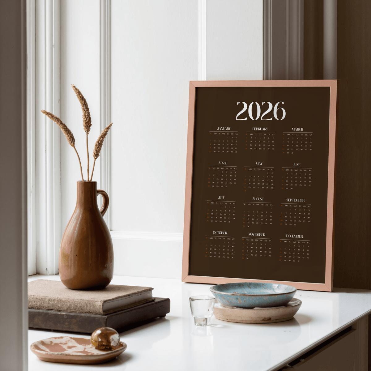 Framed Brown Calendar Print 2026 with minimalist typography leaning on a white shelf near a brown vase