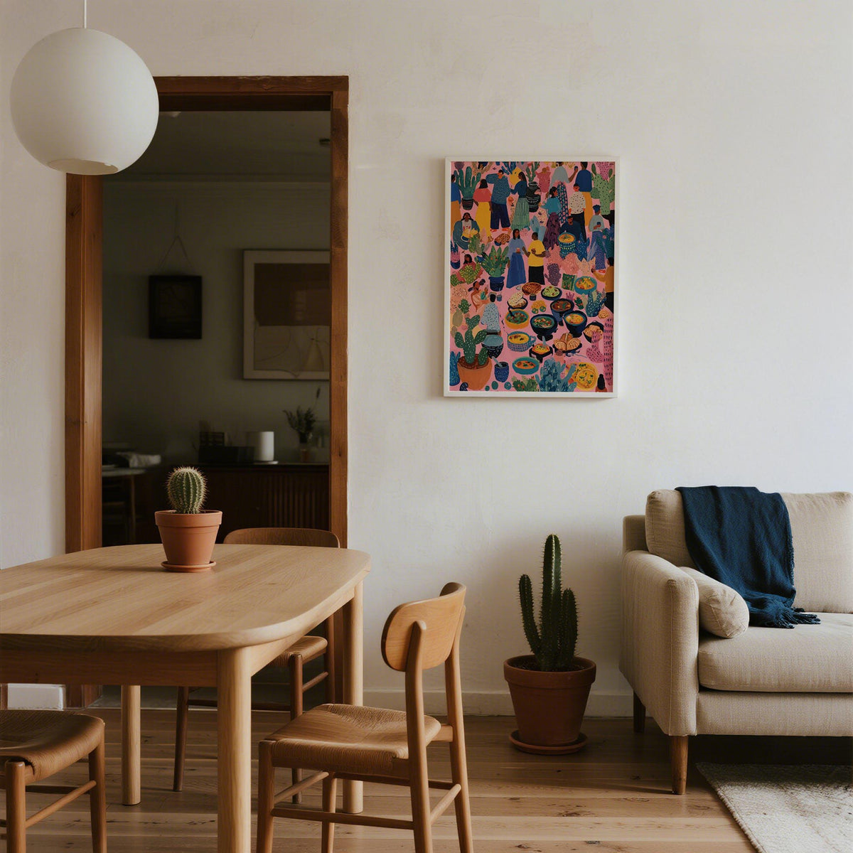 Artwork from Mexico on a white wall above a cactus in a living room with wooden furniture and a sofa