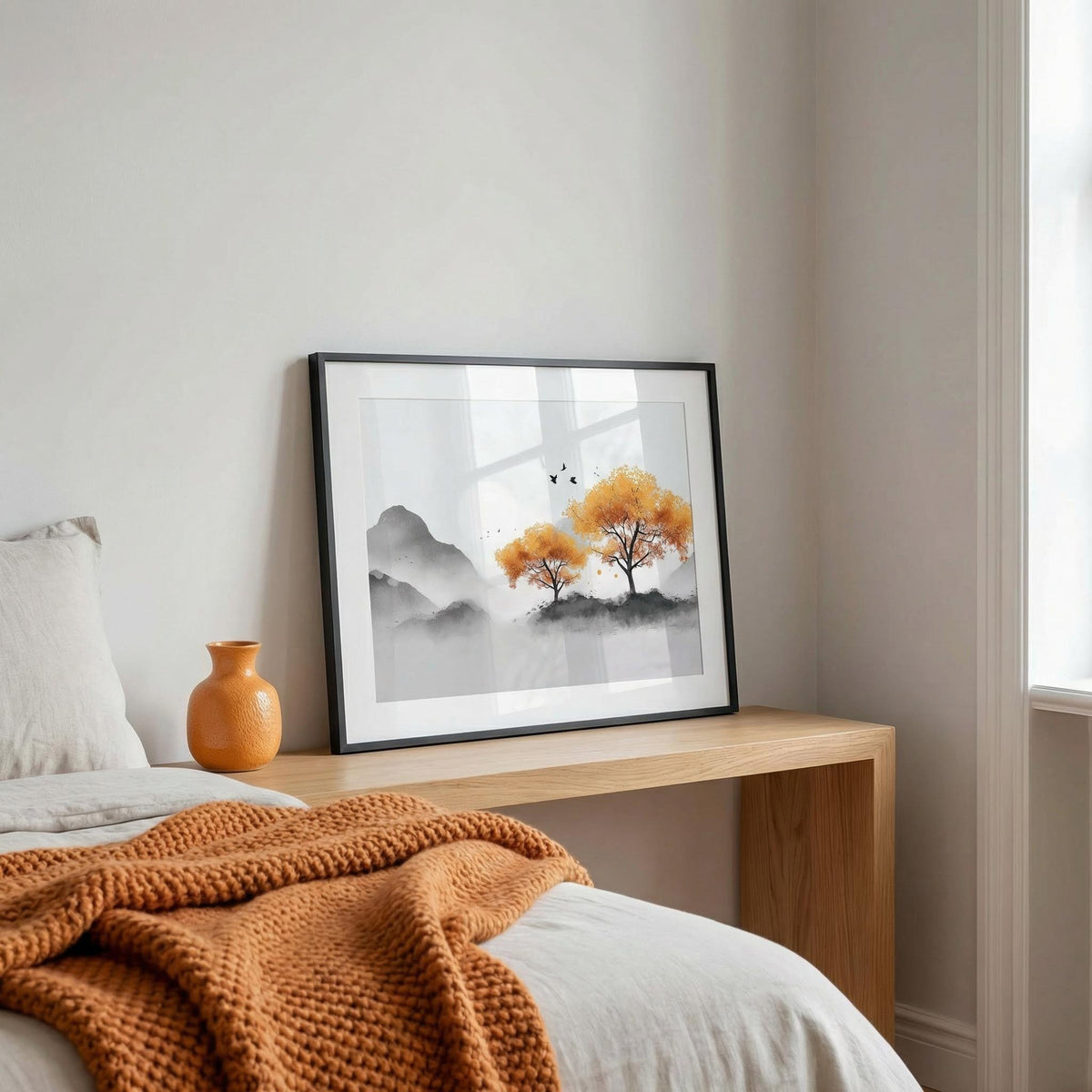 Print leaning on a wooden table of autumn tree wall art with orange leaves and grey mountains near a bed