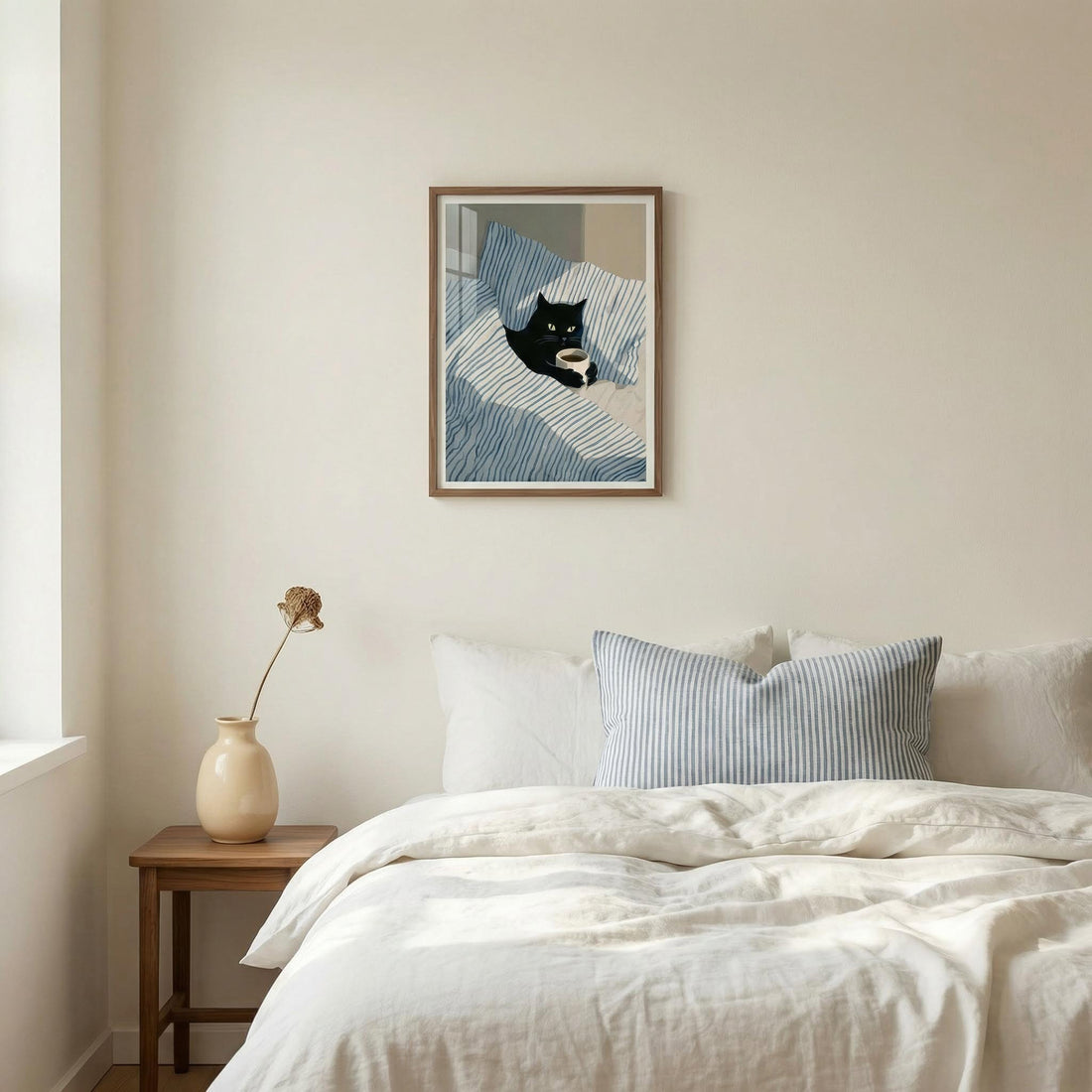 Print of a black cat reading in bed hung above a bed with white linen and a striped cushion
