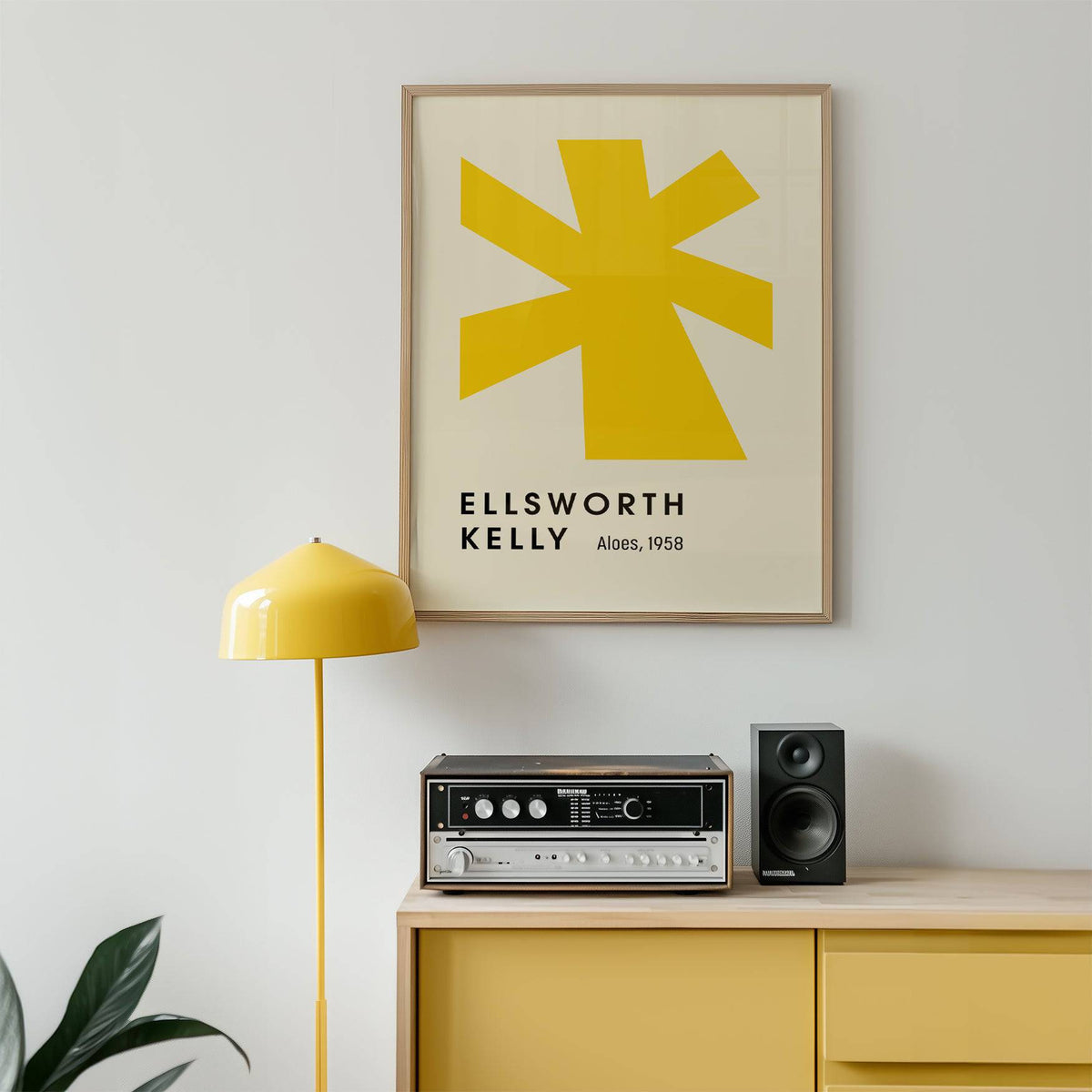 Framed print of a yellow abstract shape and black text above a wooden sideboard with a lamp and speaker