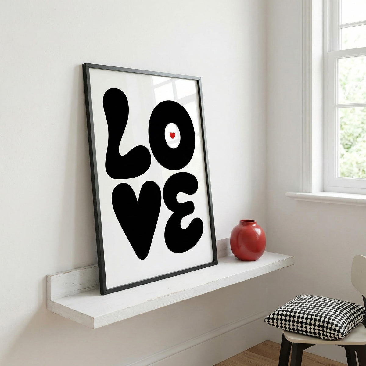 Framed close-up of Love Art Print in black and white with a small red heart leaning on a white shelf with a red vase