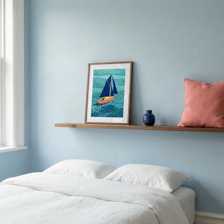Print leaning on a shelf of a colourful sailboat illustration with blue and coral hues above a bed with white pillows