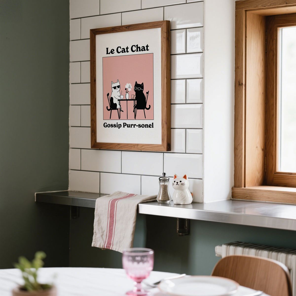 Funny Cat Artwork framed print of two cats at a table with pink background above kitchen counter with ceramic cat figurine
