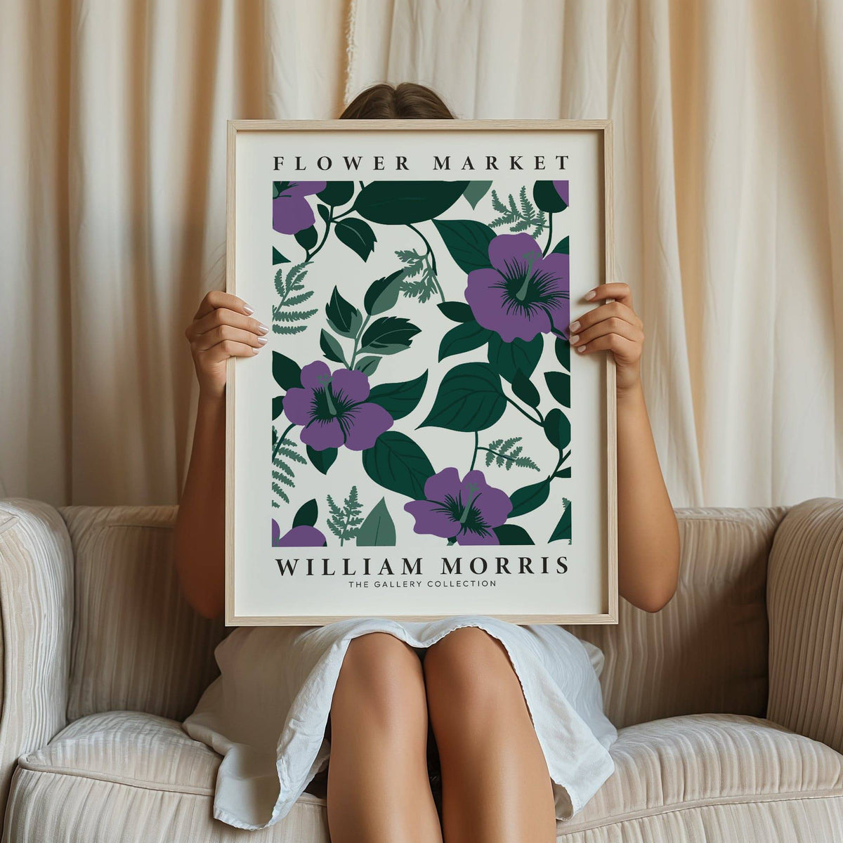 Framed purple flower artwork held by a person wearing a white dress sitting on a beige sofa