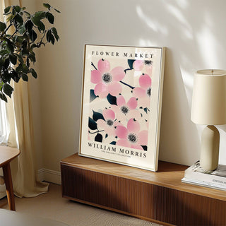 Print leaning on a wooden sideboard showing pink dogwood flowers with dark leaves beside a green plant and lamp