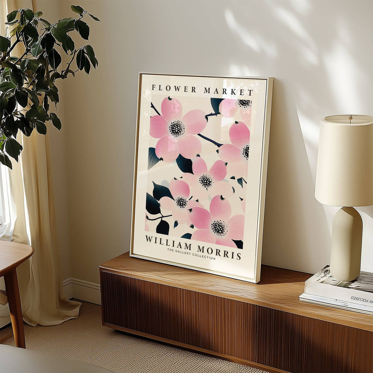 Print leaning on a wooden sideboard showing pink dogwood flowers with dark leaves beside a green plant and lamp