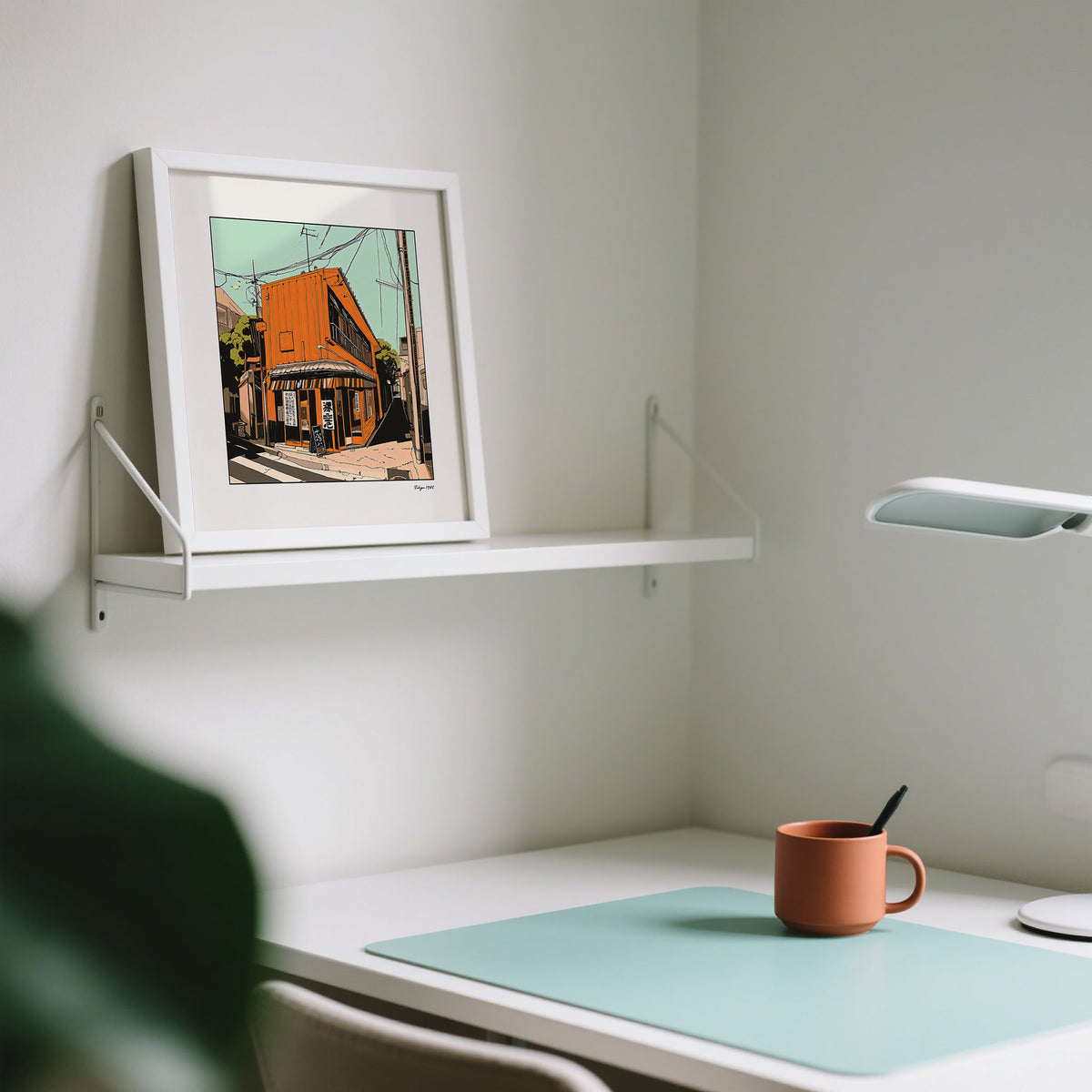 Print leaning on a white shelf showing orange building illustration in soft green and beige tones above a desk with a rust-coloured mug