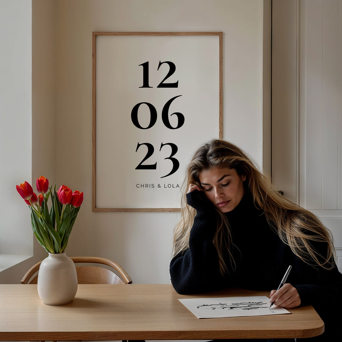 Woman writing at a table beneath a personalised date print, with a vase of red tulips beside her
