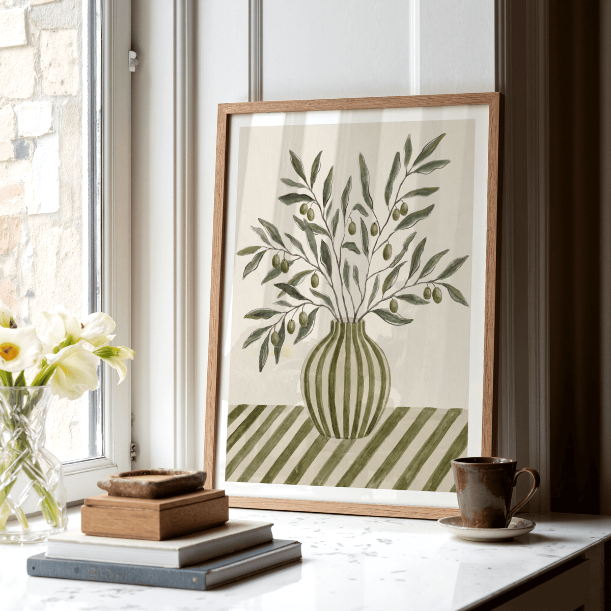 Framed Olive Branch Art print with green and brown hues leaning on a window sill beside a cup and books