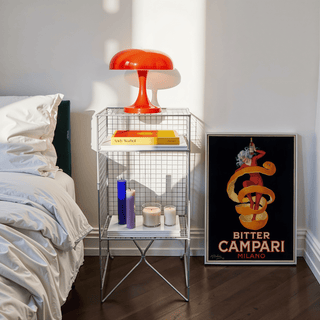 Print leaning on a floor next to a bedside table with candles and a red lamp in natural light