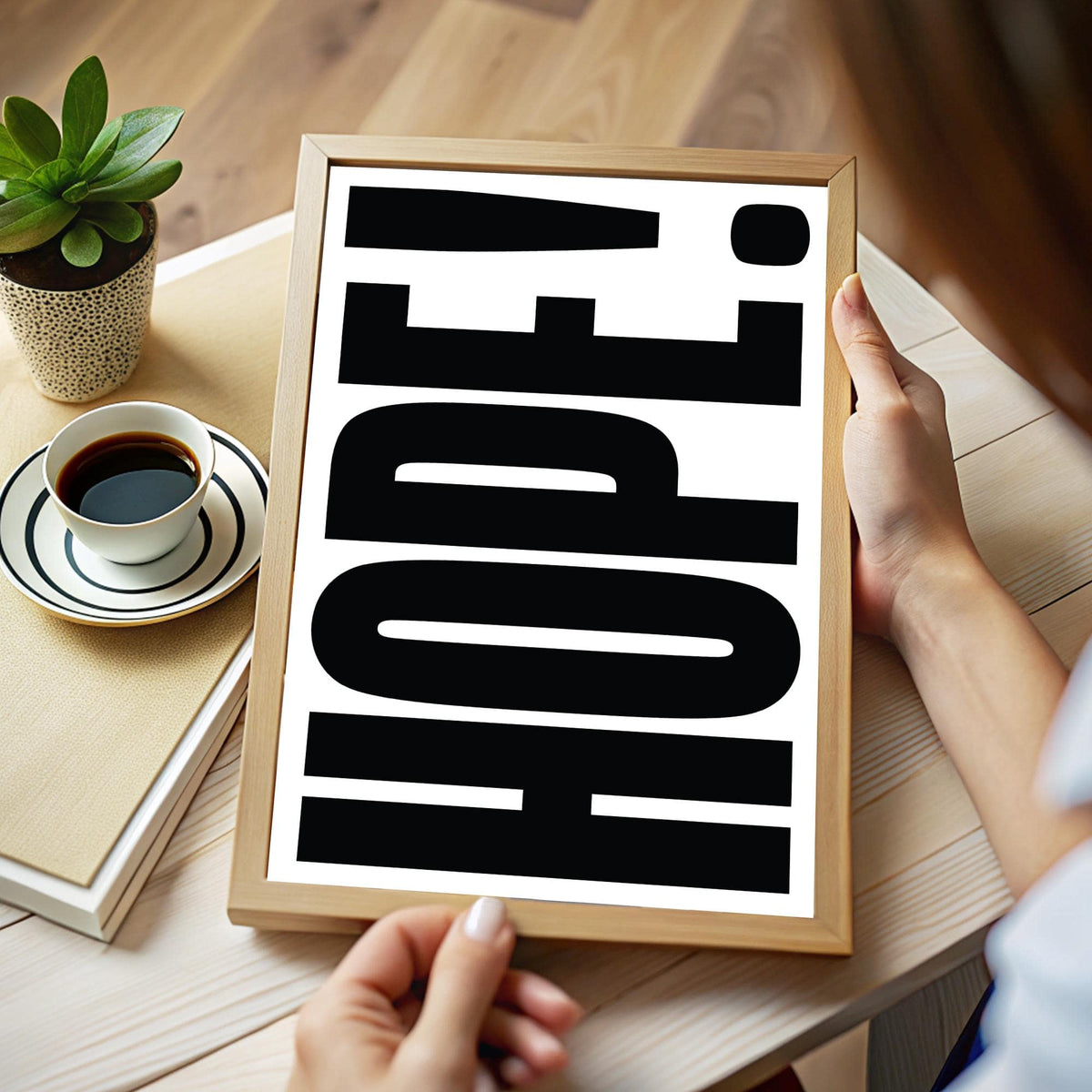 Framed HOPE typography print in black and white held in hands on a wooden table with cup and plant