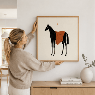 Framed Horse Moon wall art with a black horse and orange blanket held on a white wall above a wooden sideboard with books and a vase