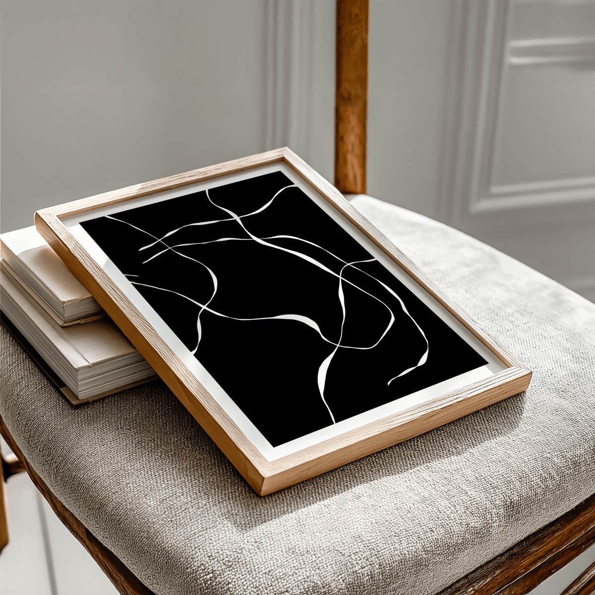 Framed Abstract Lines Art Print with white abstract lines on black leaning on a textured chair with books