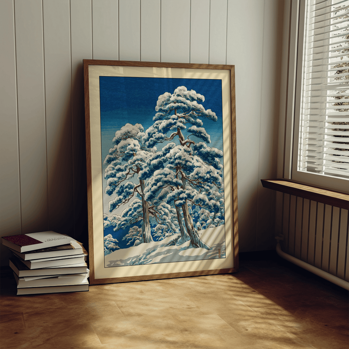 Print leaning on floor of Hasui Kawase Winter Landscape Print with snow-covered pine trees and blue sky by window with blinds