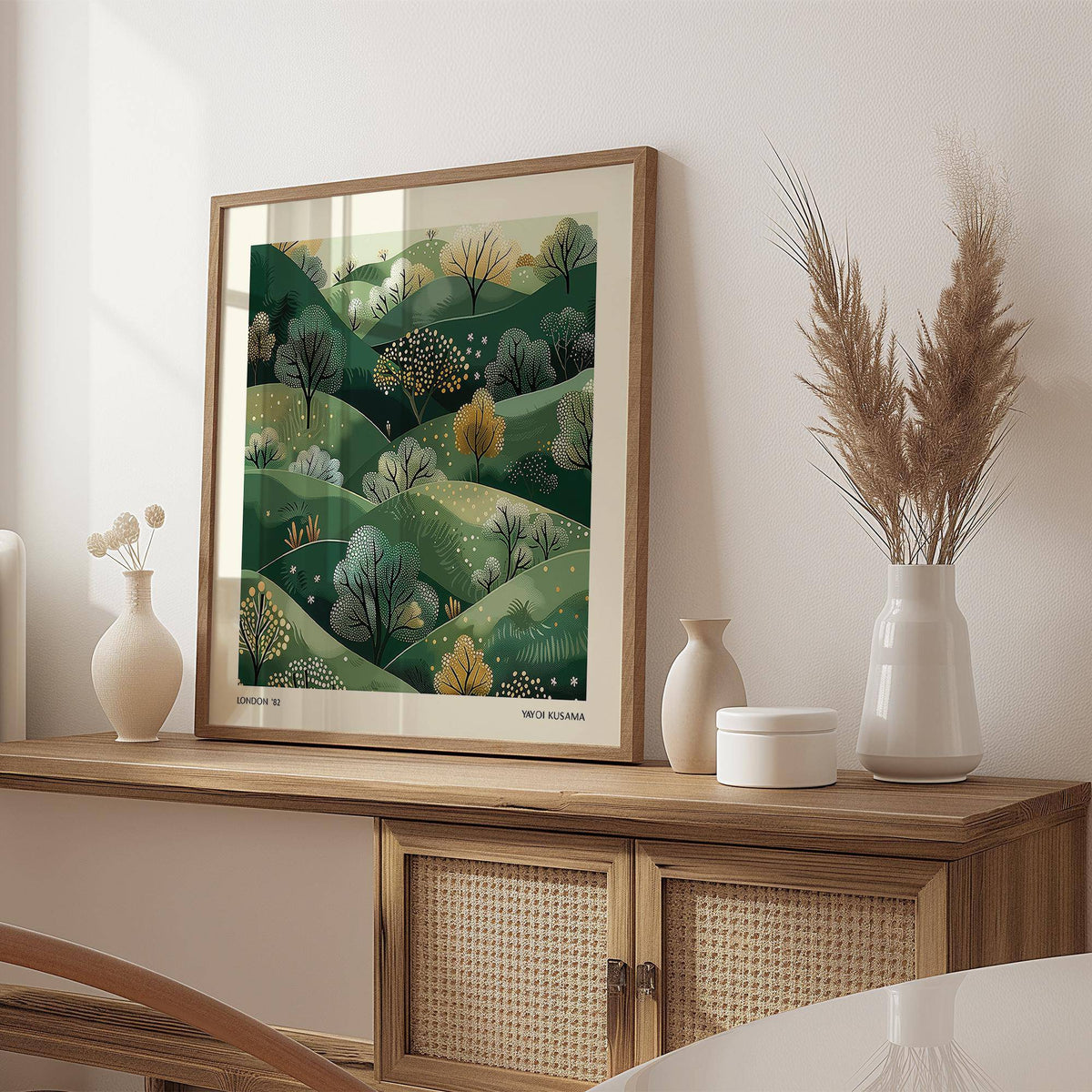 Large nature wall art of green hills with trees in an illustration style leaning on a wooden sideboard beside neutral vases