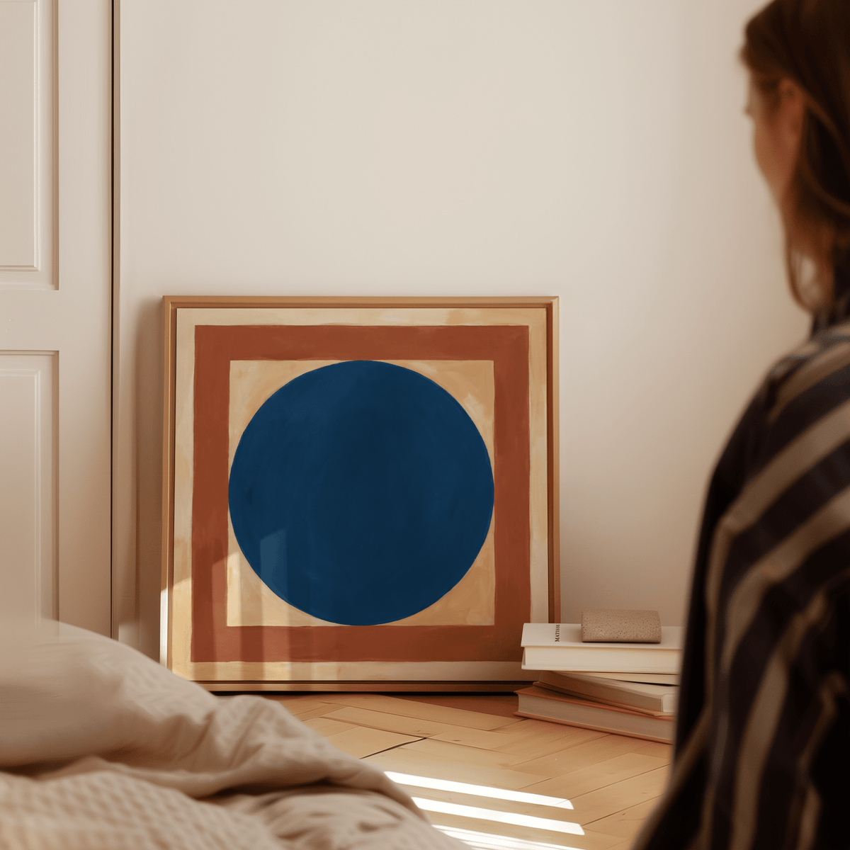 Minimalist Circle Art leaning on a floor beside stacked books with a cream wall and person nearby