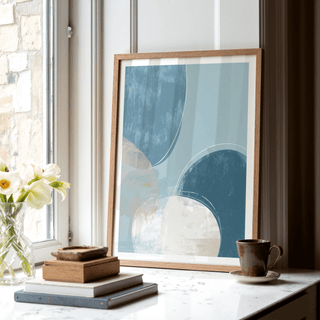 Print leaning on a white windowsill showing abstract blue circles with a brown cup and books nearby