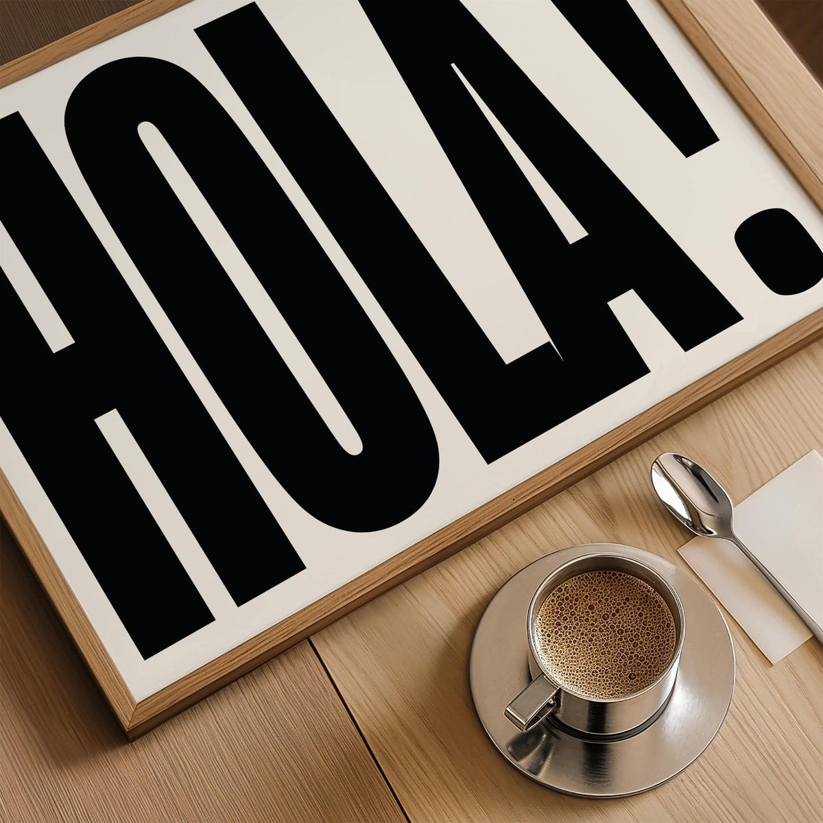 Framed close-up of typographic posters with bold black HOLA text and light wood frame on a table with a cup of coffee